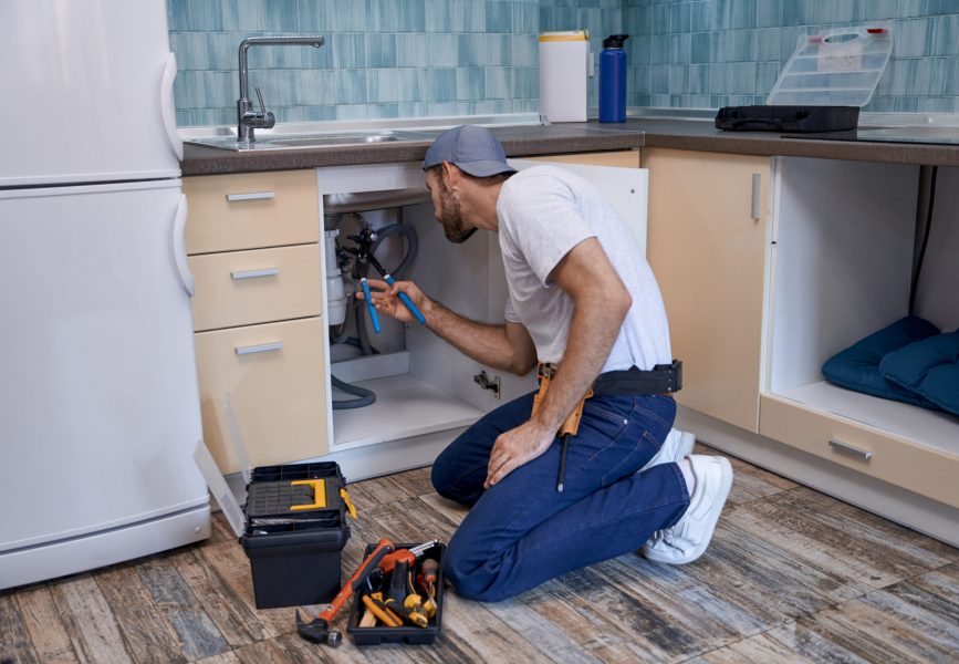 Young caucasian male worker using adjustable wrench while fixing tubes under kitchen sink looking into drawer. Home master concept, construction and repair